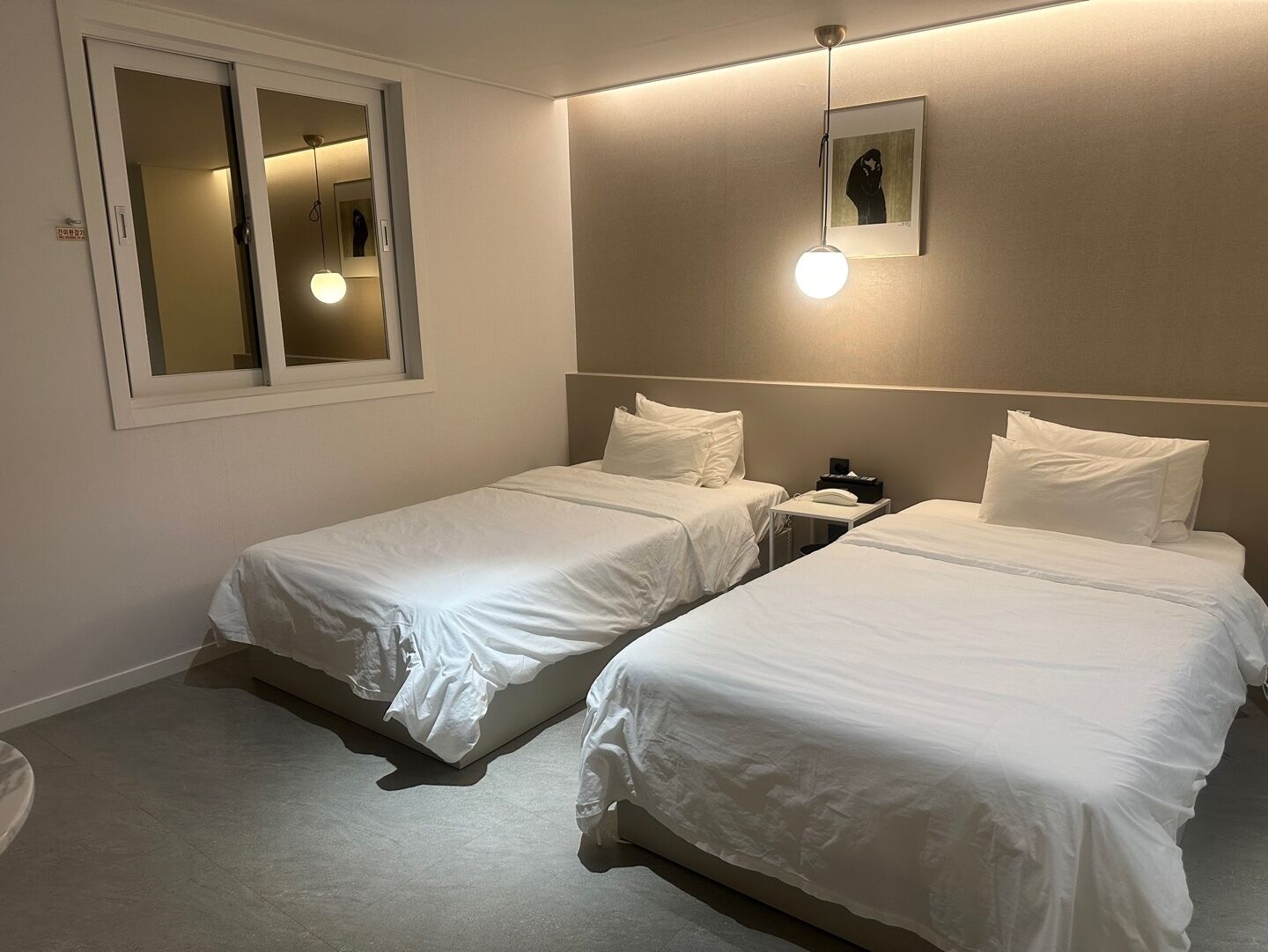 A hotel room with two single beds, a bedside table with a lamp, a small bedside table with a phone, and a framed picture on the wall.
