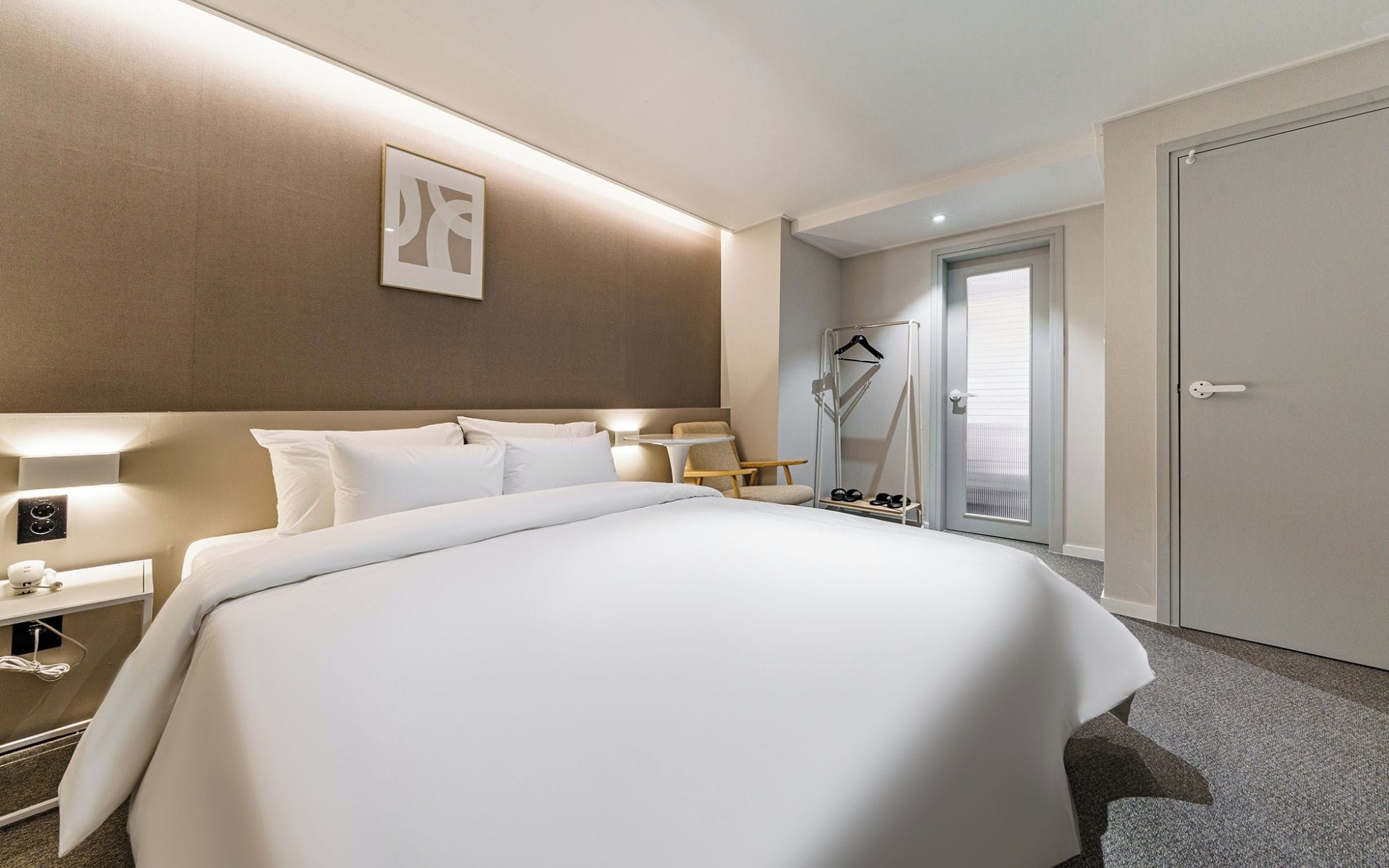 Quarto de hotel com uma cama grande, mesas de cabeceira, uma cadeira, um cabideiro e uma porta.