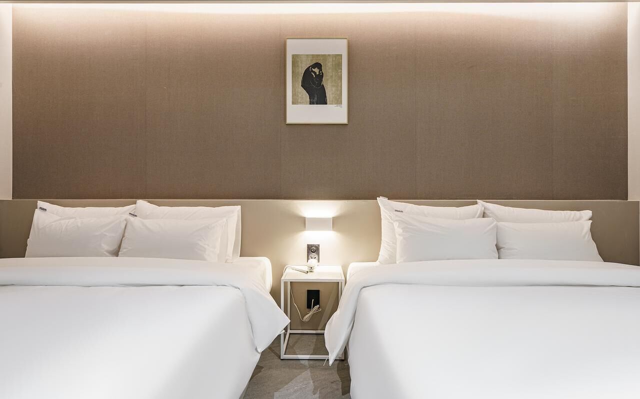 A hotel room with two beds, a bedside table with a lamp, and a framed picture on the wall.