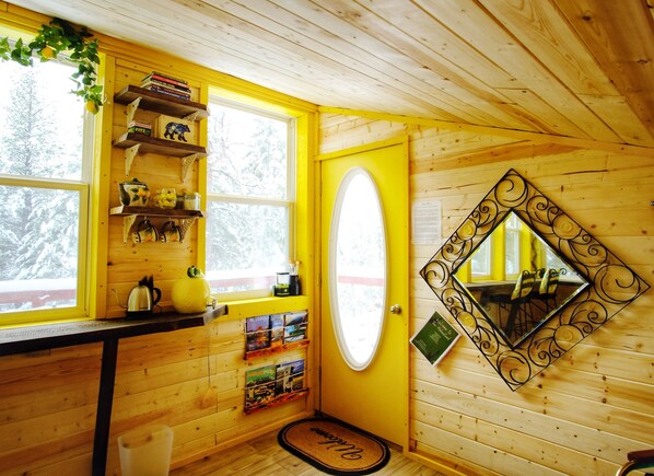 Ein gemütliches Blockhaus-Interieur mit Holzwänden, einer gelben Tür, einem Fenster mit Blick auf schneebedeckte Bäume, Regalen mit Büchern und Küchenutensilien sowie einem dekorativen Spiegel.