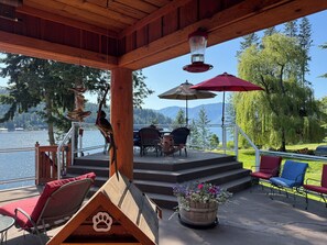 A covered outdoor seating area with a wooden deck, a doghouse-shaped planter with flowers, and a view of a lake and mountains. - LAKESIDE LODGE. GARFIELD BAY WATERFRONT.  TWO DECKS. DOCK. GREAT MOUNTAIN VIEWS. (Sagle)