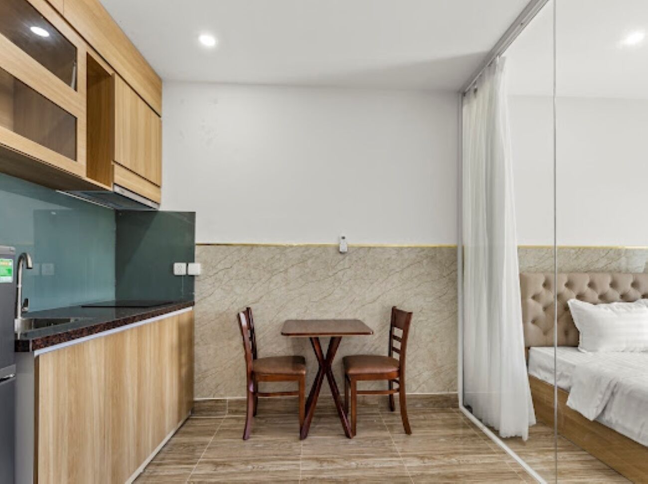 A compact room with a kitchenette, a dining table with chairs, a bed with curtains, and a marble accent wall.