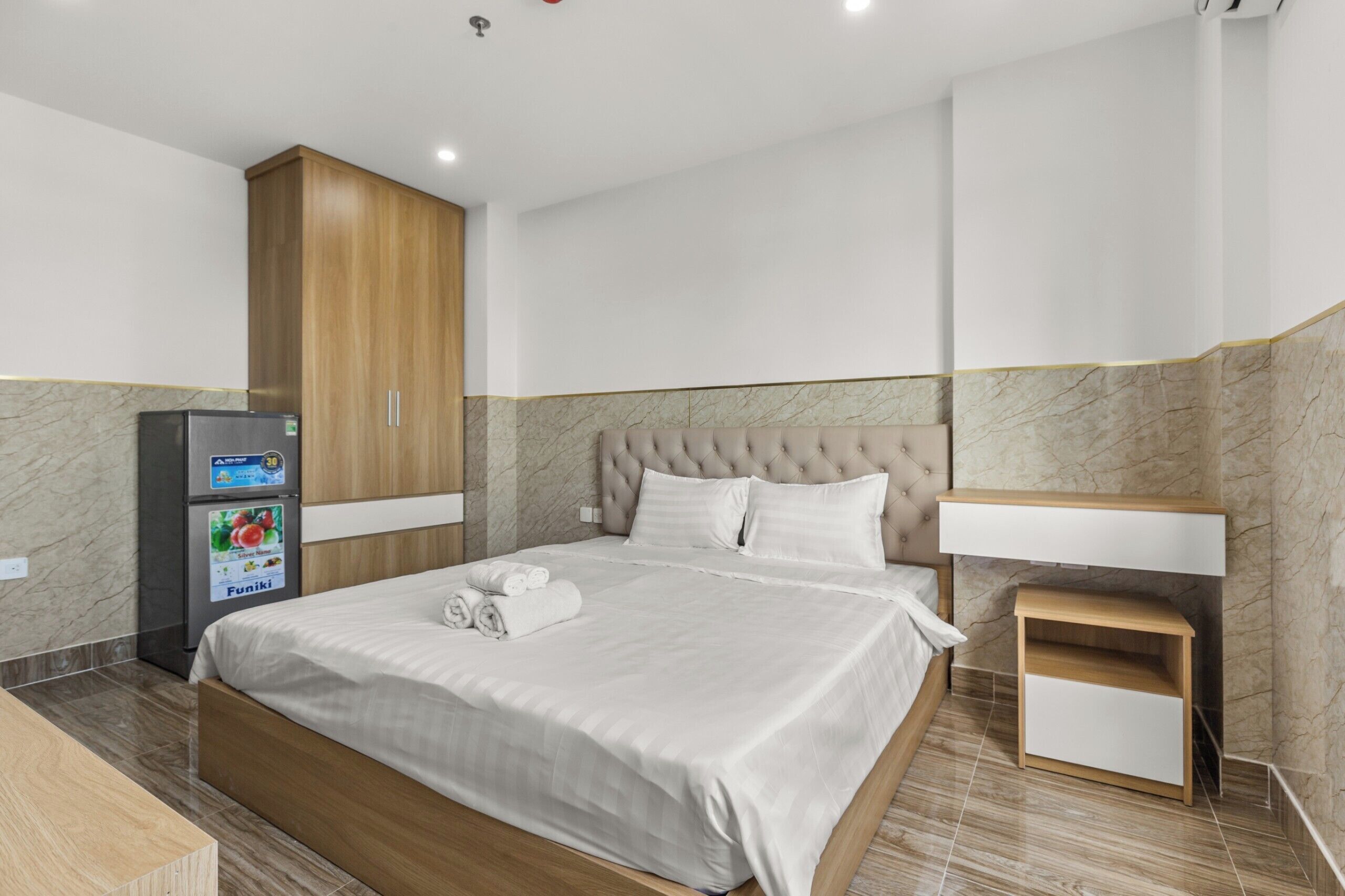 A hotel room with a bed, a minibar, a wooden wardrobe, a bedside table, and a marble-patterned wall.