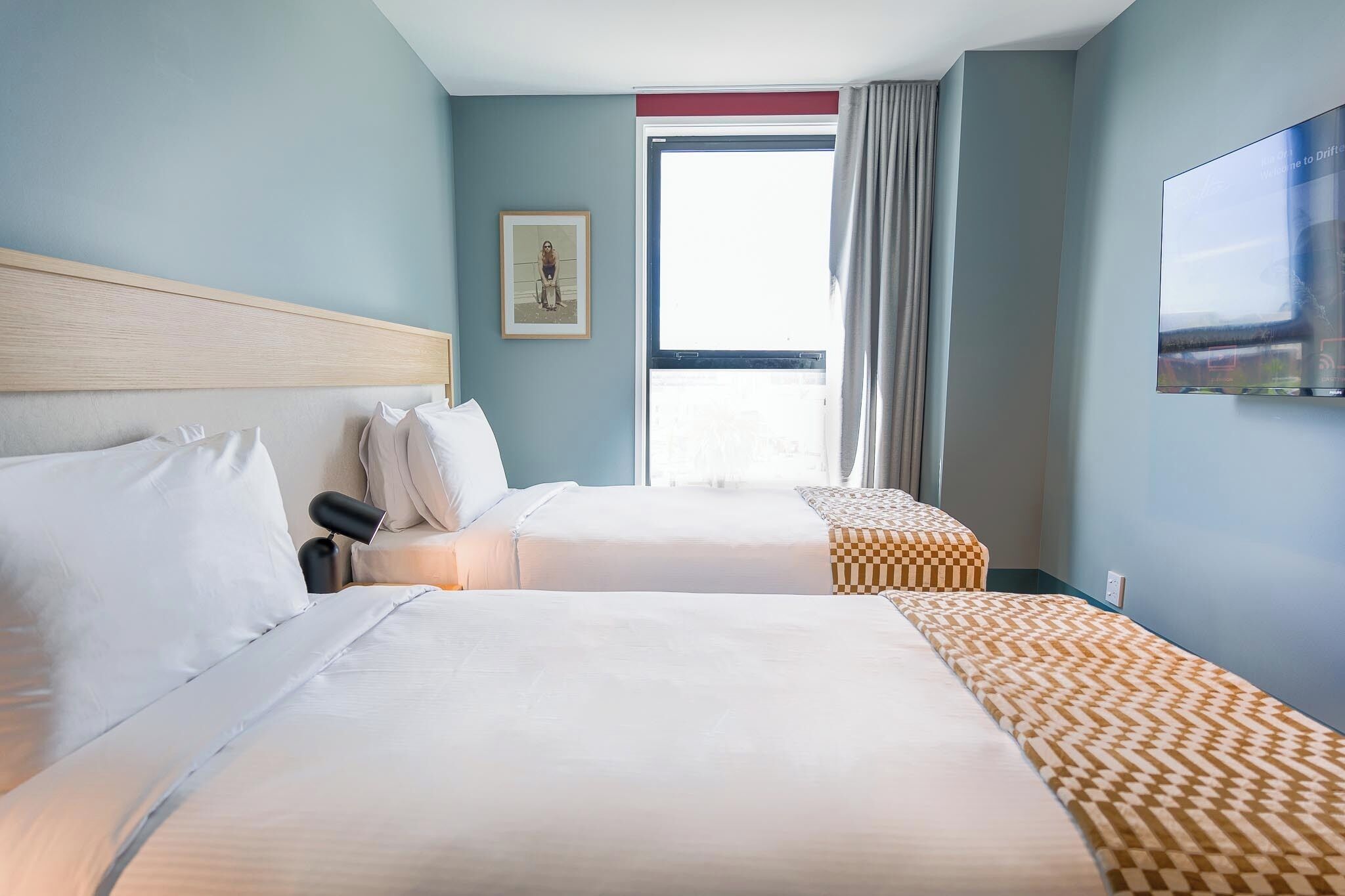 A hotel room with two beds, a large window with curtains, a mounted TV, and a framed picture on the wall.