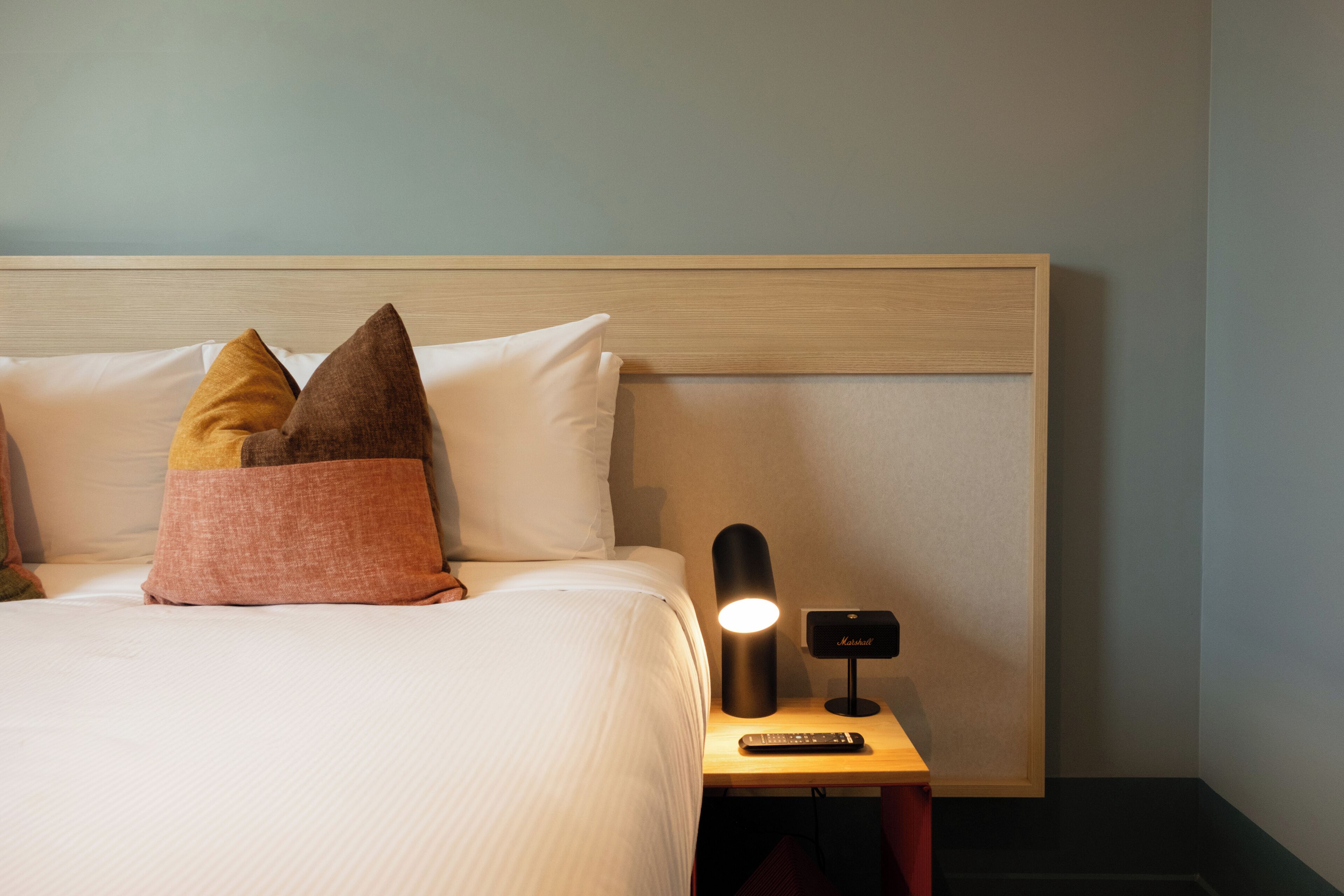 Een opgemaakt bed met kussens, een bedlampje en een afstandsbediening op een nachtkastje van hout.