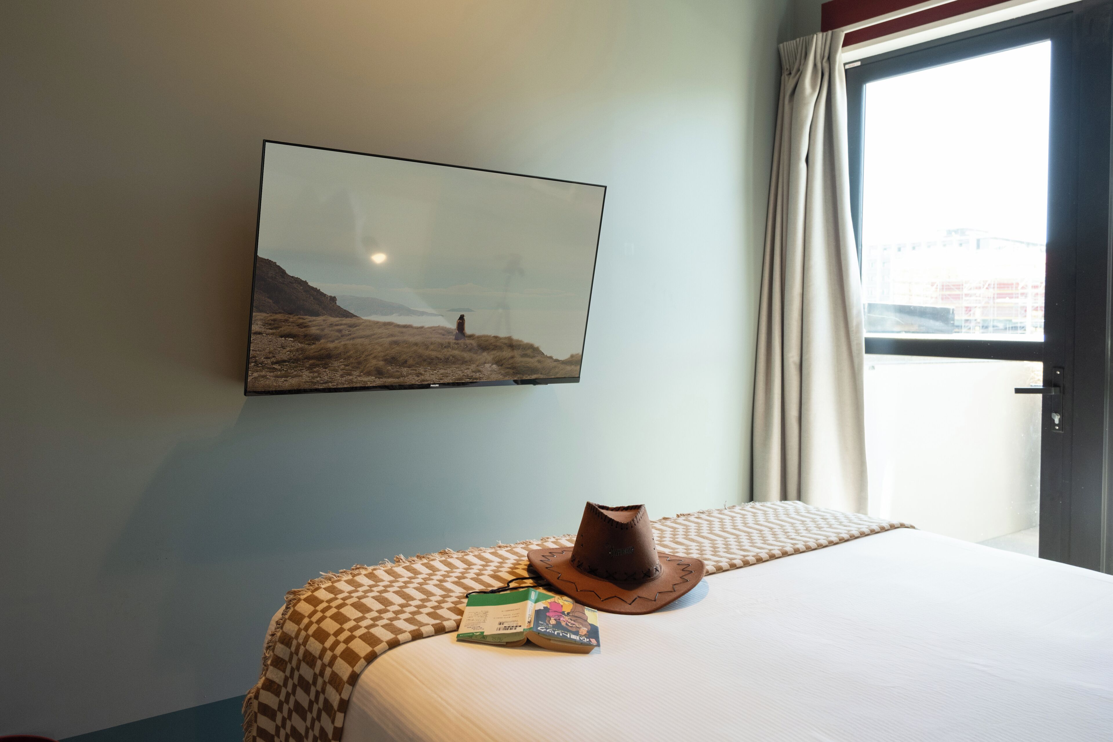 Ein Hotelzimmer mit einem Bett, einer karierten Decke, einem Hut und einem gerahmten Landschaftsbild an der Wand.