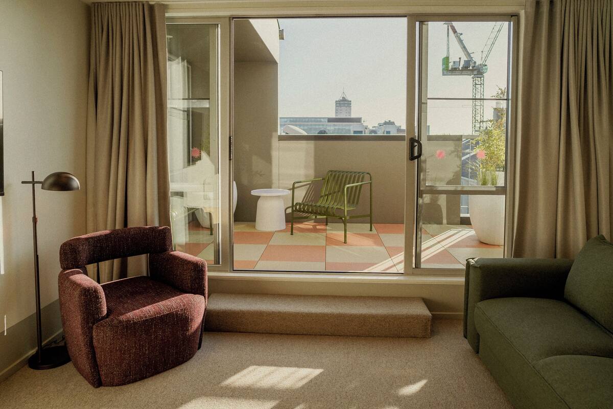 Una sala de estar con un sofá verde, un sillón rojo y una silla verde junto a un gran ventanal corredera.