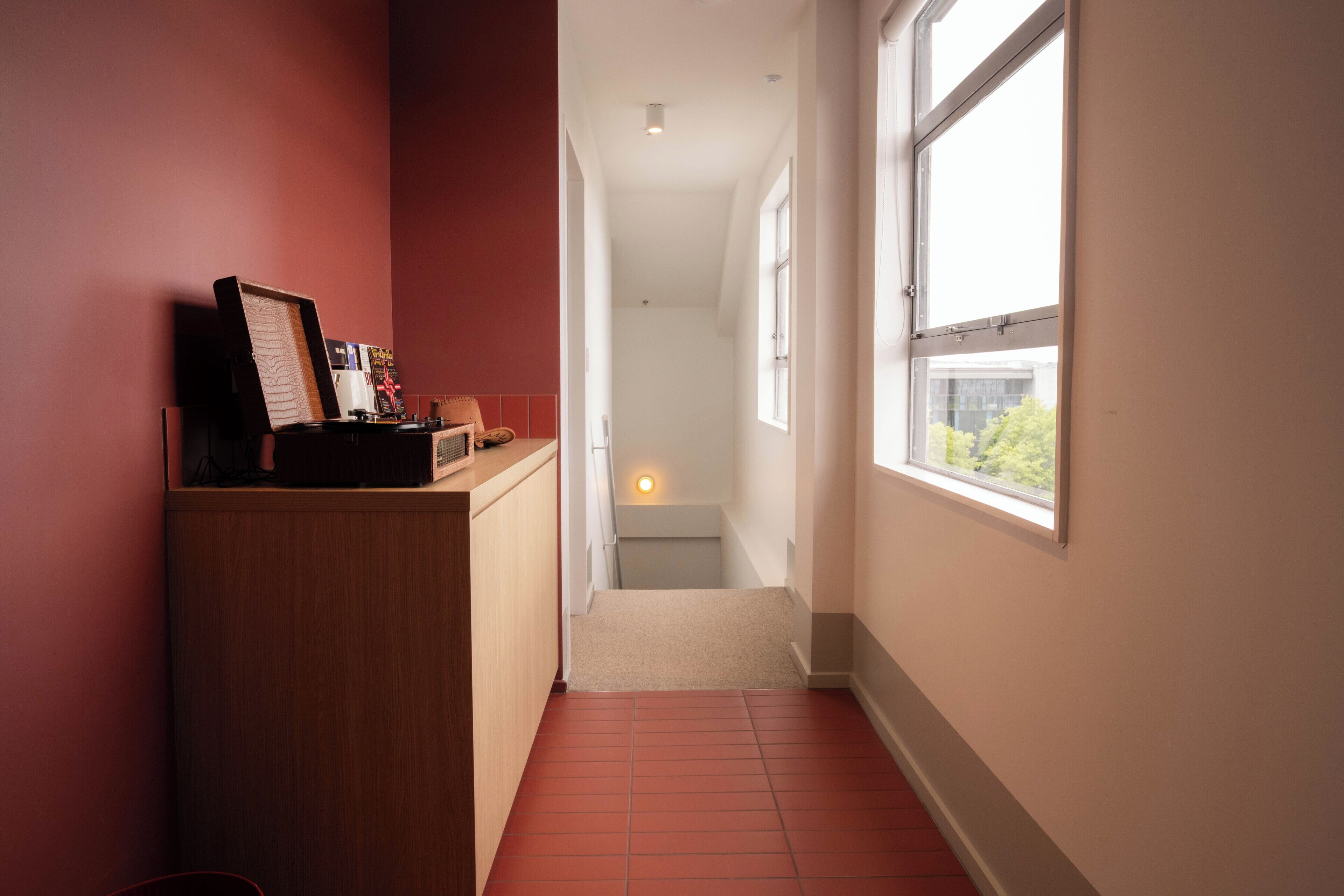 A narrow hallway with a red-tiled floor, a wooden cabinet with a picture frame, and a window on the right.
