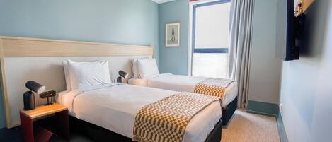 A hotel room with two beds, a desk with a lamp, a TV mounted on the wall, and a window with curtains.