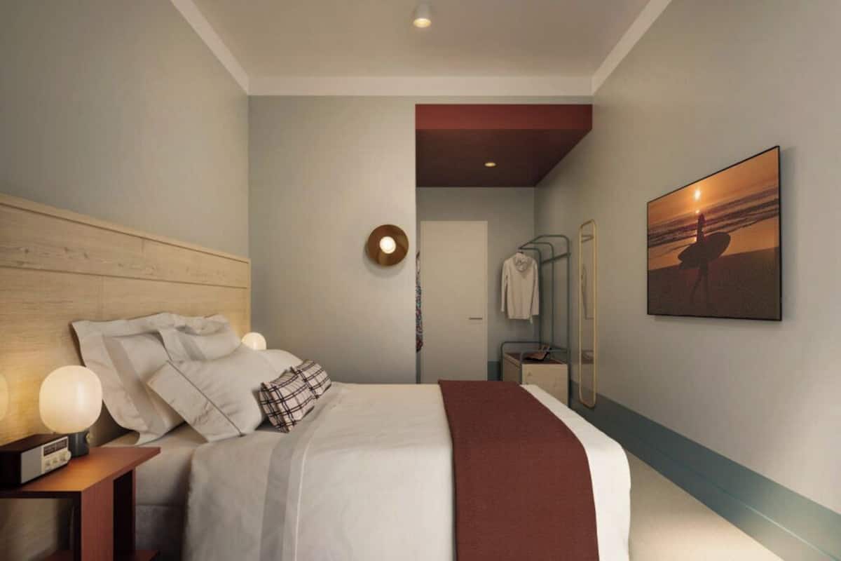 Habitación de hotel moderna con cabecera de madera, una cama grande con ropa de cama blanca y un plaid marrón, una mesita de noche con lámpara, un espejo montado en la pared y un cuadro con una imagen del atardecer.