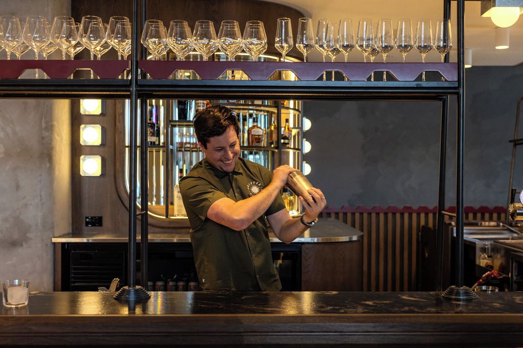 A bartender preparing drinks behind a bar with a wine rack above and a well-lit interior.
