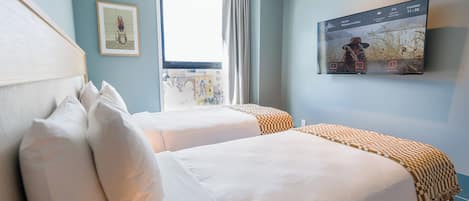 A hotel room with two beds, a large window, a mounted TV, and a framed picture on the wall.