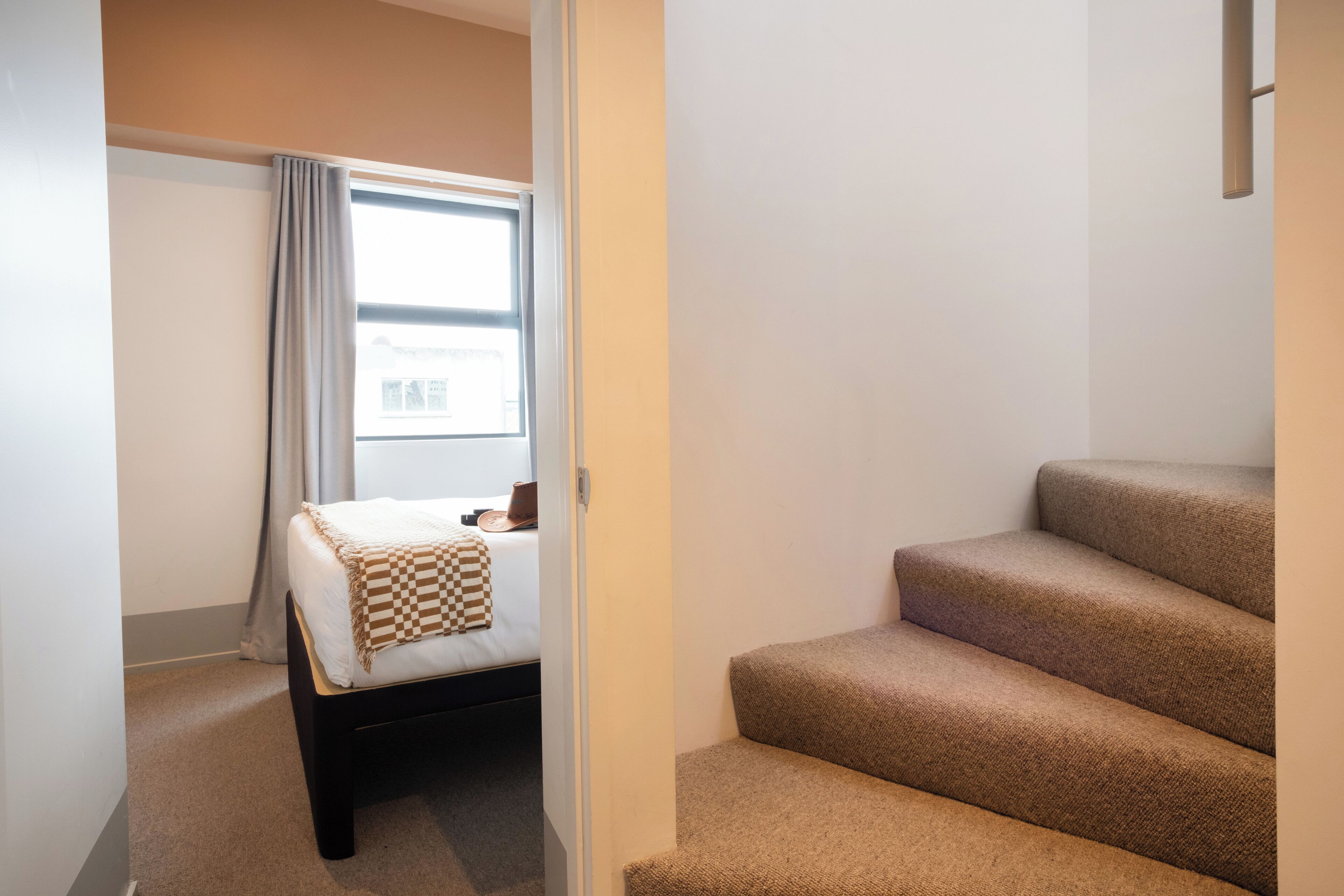 A small, compact room with a bed, a window with curtains, and a carpeted staircase.
