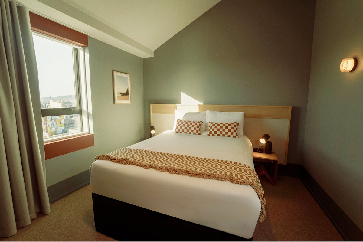 Habitación de hotel con una cama grande, una ventana con cortinas, una mesita de noche con lámpara y una luz en la pared.
