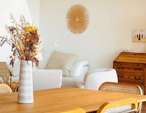 Un coin repas avec une table en bois, des chaises blanches, un vase contenant des fleurs séchées, un canapé blanc, une commode en bois et une œuvre d’art murale.