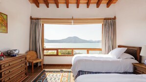 Un dormitorio con dos camas, un aparador de madera, una silla, un ventanal con vistas a un lago y montañas, y una alfombra estampada.