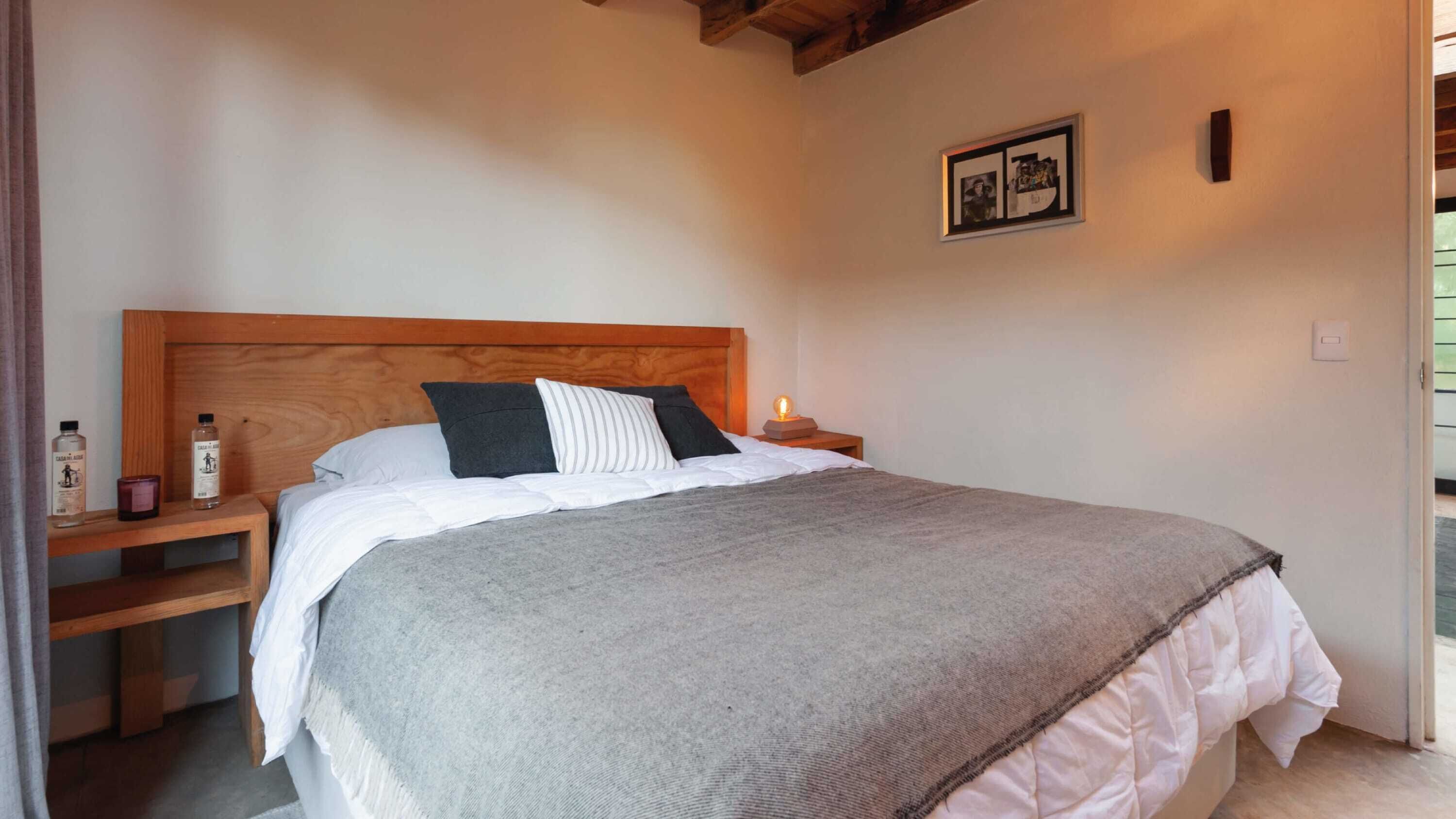 Una cama bien hecha con cabecera de madera, una mesita de noche con lámpara y un cuadro enmarcado en la pared.