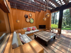 Un intérieur chaleureux avec un canapé, une table basse et un téléviseur. Des lampes suspendues illuminent l’espace et, par la fenêtre, on peut admirer la vue sur les arbres.