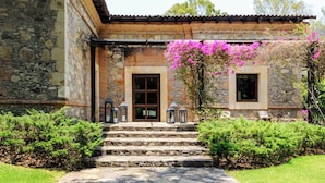 A stone building with a tiled roof, a set of stone steps leading to a door, and a garden with green shrubs and pink flowers. - El Salto: Luxury Ranch with Lake View (Valle de Bravo)