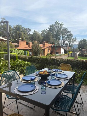 An outdoor dining area with a table set for four, surrounded by green chairs, and a bowl of fruit on the table.