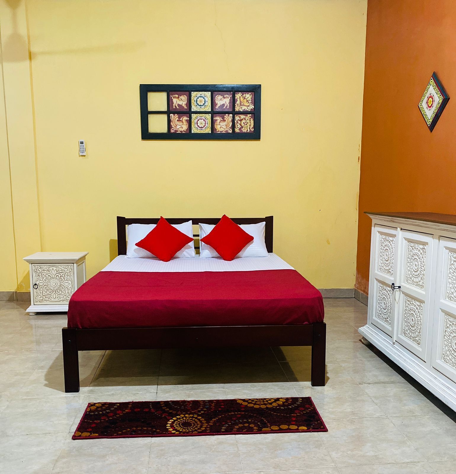 Ein Bett mit roter Bettdecke und zwei roten Kissen, ein gemustertes Teppich, eine weiße Kommode mit aufwendigen Verzierungen und eine dekorative Wandverkleidung.