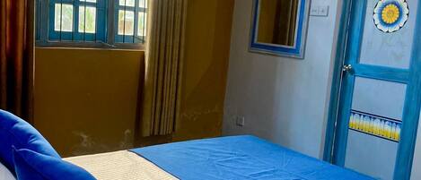 A room with a bed, a window with curtains, a door with a decorative panel, and a wall-mounted mirror.