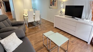 Un salon moderne avec un canapé gris, une petite table blanche, un téléviseur posé sur un meuble blanc, et un coin repas à l’arrière-plan.