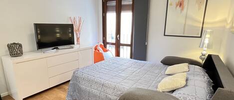A bedroom with a bed, a TV on a white dresser, a grey sofa, and a lamp.