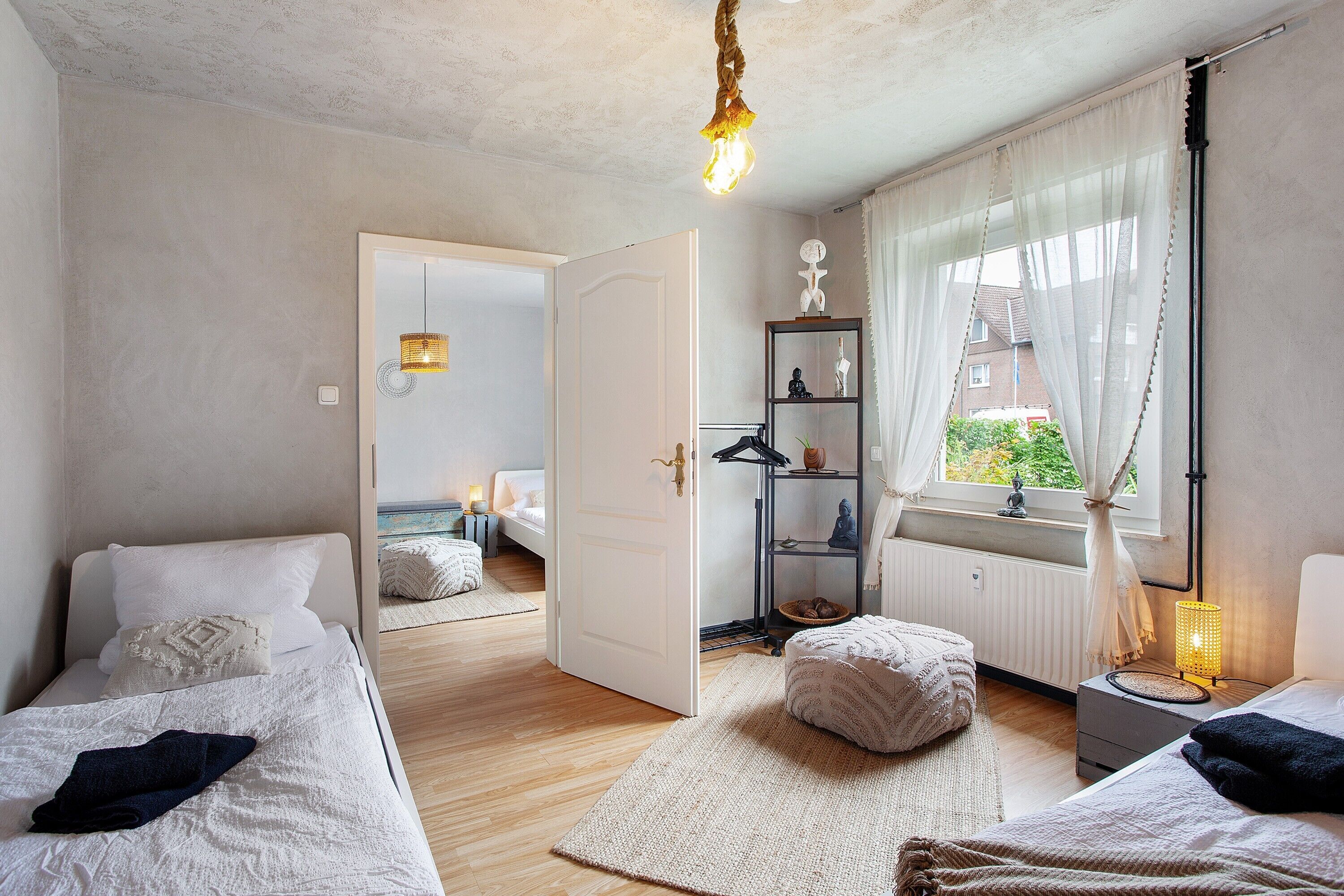 A bedroom with two beds, a bedside table with a lamp, a window with curtains, and a door leading to another room.