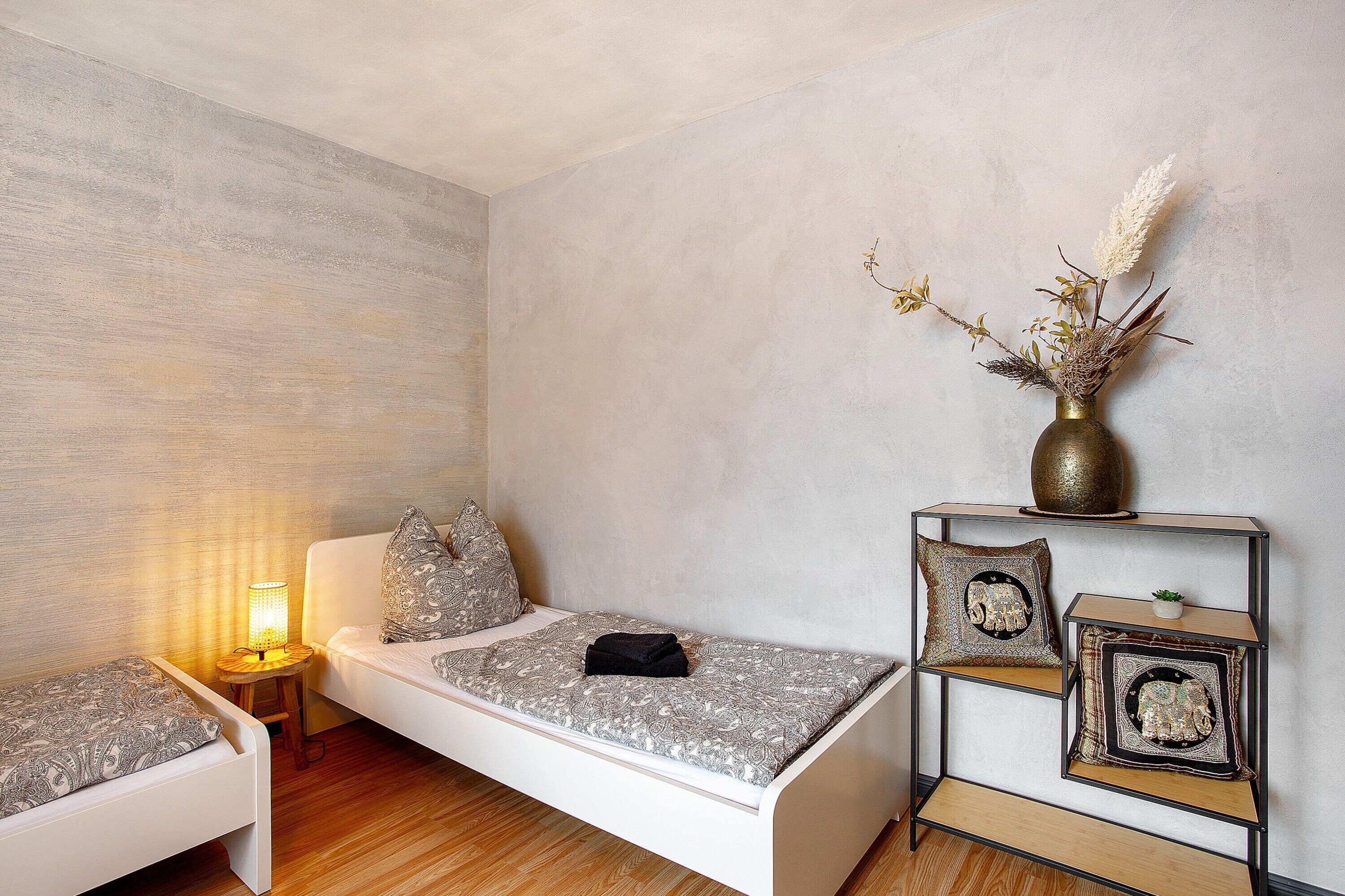 A bedroom with a single bed, bedside table, lamp, and decorative items including a vase with flowers.