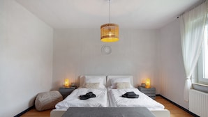 Ein Schlafzimmer mit zwei Betten, Nachttischlampen, einer Hängelampe, einer Wandplatte zur Dekoration und einem Fenster mit Vorhängen.