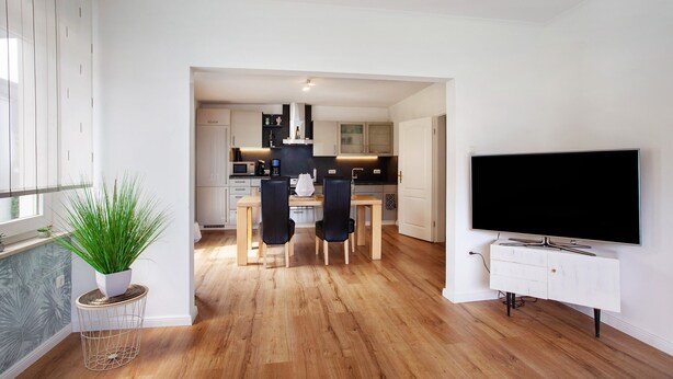 Eine moderne Küche mit Holzboden, einem Essbereich mit Stühlen, einer Topfpflanze und einem Fernseher auf einem weißen Schrank.