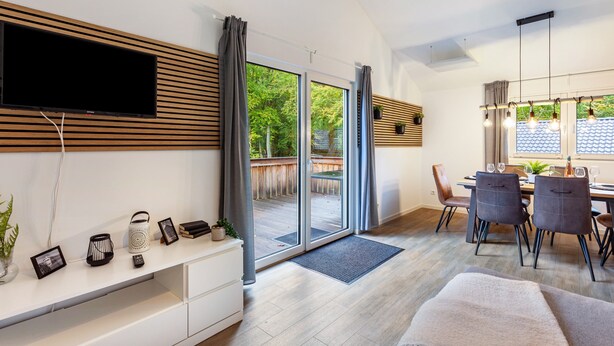 A modern living room with a flat-screen TV, a dining area with chairs and a table, and a sliding glass door leading to an outdoor deck. - Vacation Home "14" with Private Terrace & Wi-Fi (Bad Arolsen)