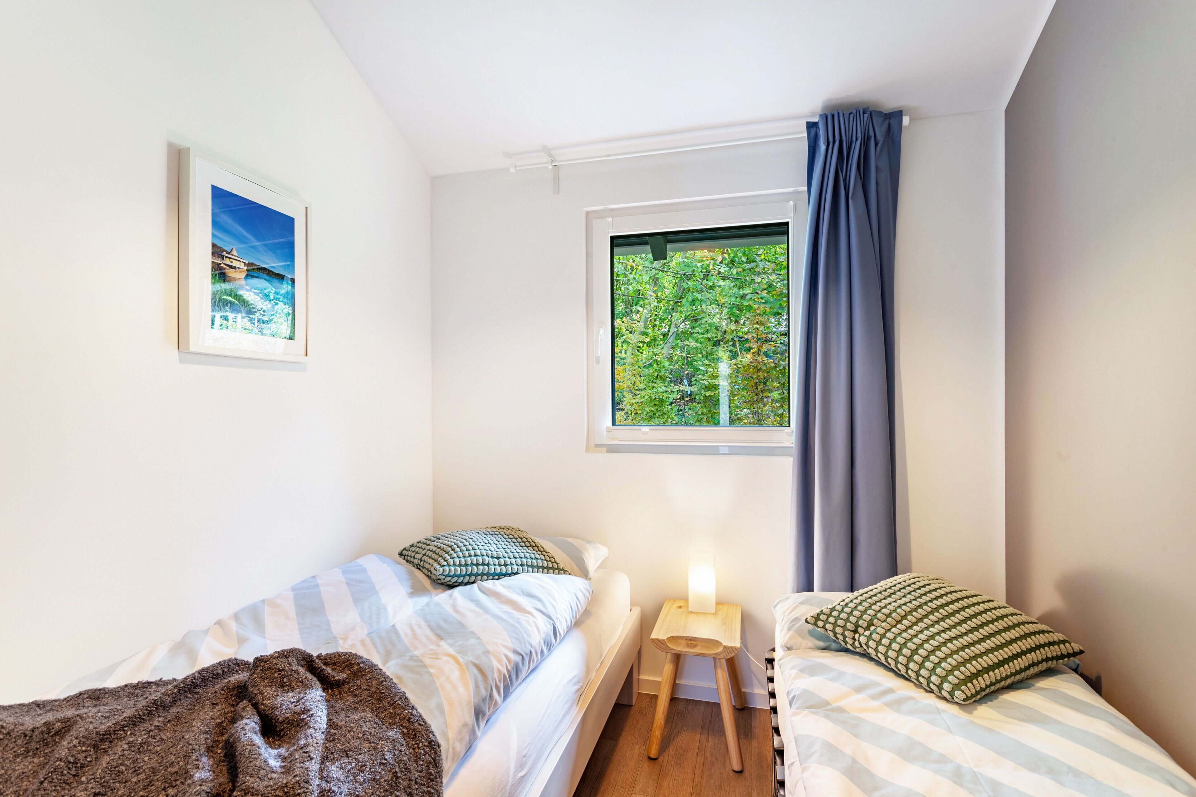 A bedroom with two beds, a window with a view of trees, a framed picture on the wall, and a bedside table with a lamp.
