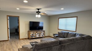 A living room with a gray sectional sofa, a wooden entertainment center, a ceiling fan, and a large window with blinds.