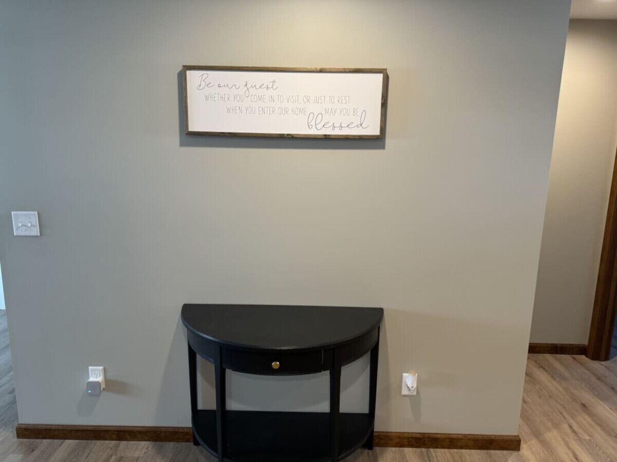 A room with a black console table against a light-colored wall, a framed sign above it, and a light switch on the wall.