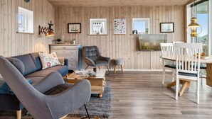 Un salon chaleureux avec un plafond en bois, un canapé, un fauteuil gris, une table à manger avec des chaises blanches et un téléviseur fixé au mur.