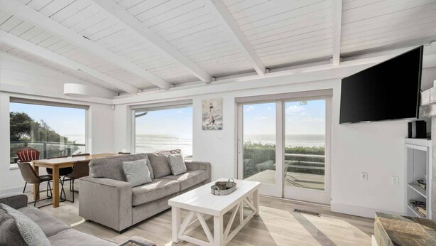 Amplia sala de estar con un sofá gris, una mesa de centro blanca y una mesa de comedor con sillas. Ventanas grandes ofrecen vistas al exterior.