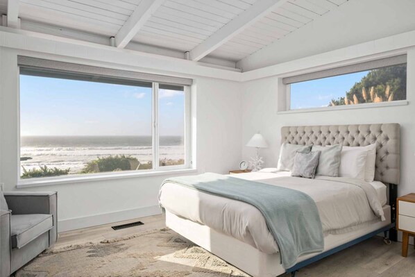 A bedroom with a large bed, two windows with ocean views, a bedside table with a lamp, and a small chair.
