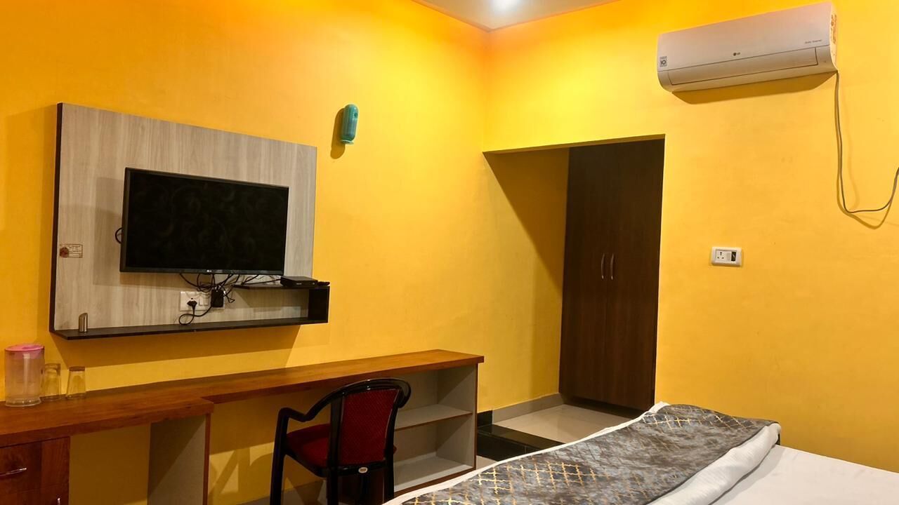 A room with a bed, a desk with a chair, a television mounted on the wall, and an air conditioning unit.
