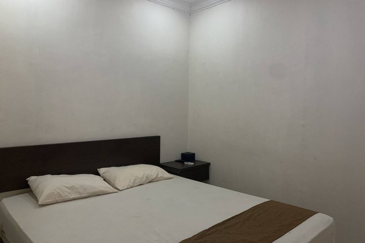 Una cama individual con ropa de cama blanca y una manta marrón, un cabecero oscuro y una mesita de noche con un objeto azul encima.