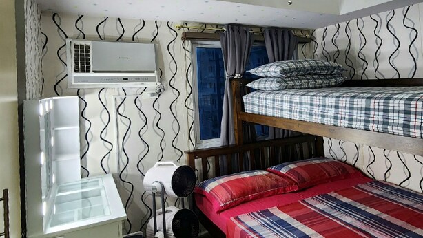 A compact room with bunk beds, a desk with a chair, a white refrigerator, a heater, and an air conditioning unit. - High-Tech Studio @ Grass Residences -2persons only (Quezon City)