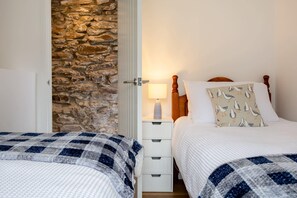 Une chambre avec deux lits, un mur en pierre, une tête de lit en bois, une table de chevet avec une lampe et un coussin à motifs.
