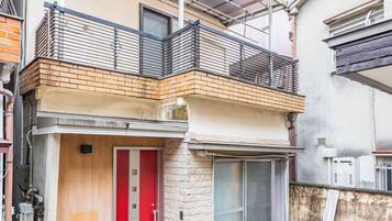 A two-story residential building with a red door, a balcony, and a small outdoor area with a concrete floor.