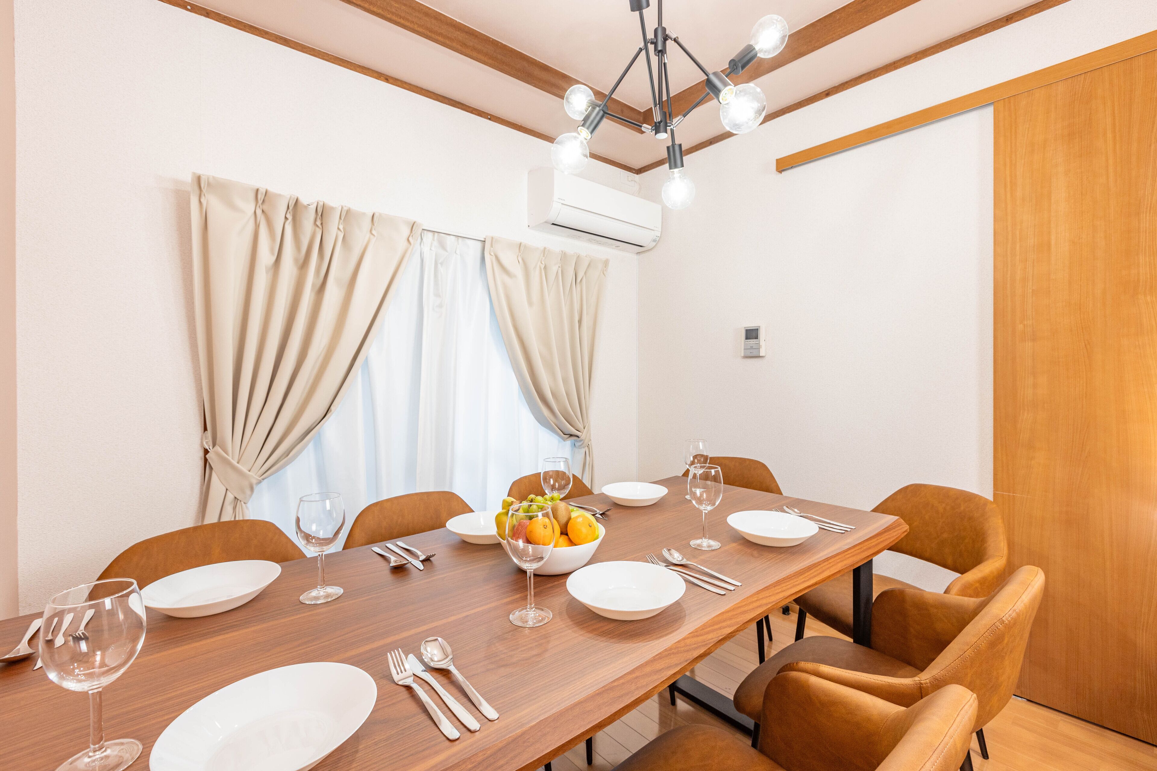 A dining area with a long wooden table set for four, including plates, cutlery, and glasses. There is a fruit bowl on the table, and the room has wooden ceiling beams and an air conditioning unit on the wall.