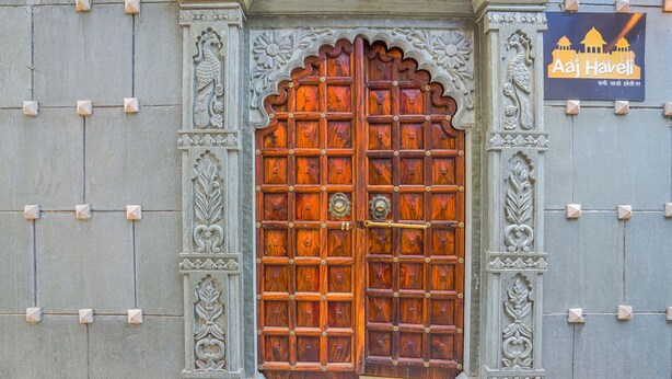 A large wooden door with intricate carvings, set within a stone wall with decorative elements. - Aaj Haveli by Levelup (Udaipur)