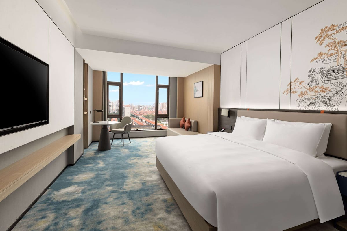 Een moderne hotelkamer met een groot bed, een flatscreen televisie, een bureau met stoel en uitzicht op de stad door grote ramen.