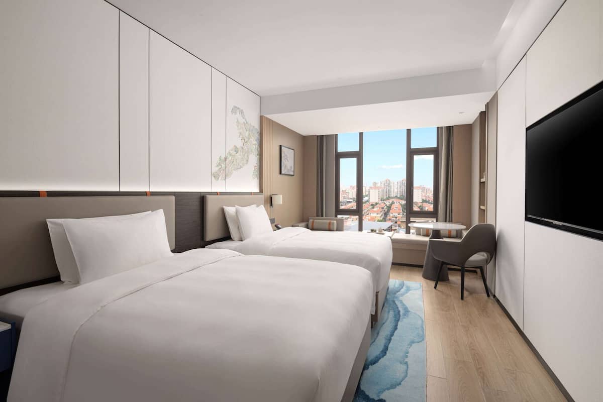 Een hotelkamer met twee bedden, een bureau, een stoel, een televisie en een groot raam met uitzicht op de stad.