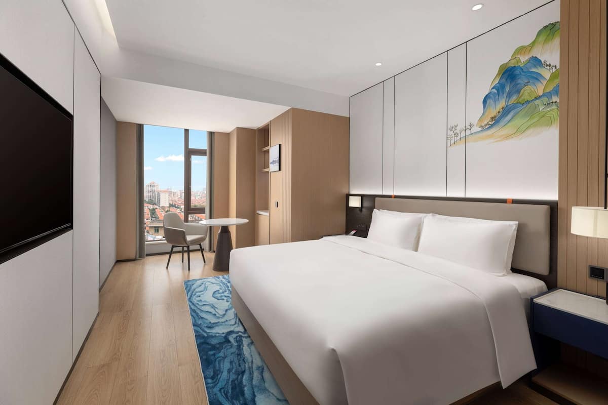 Een moderne hotelkamer met een groot bed, een bureau, een stoel en uitzicht op de stad door een groot raam.