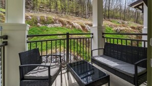 Un espace extérieur aménagé, abrité et doté de meubles en osier et d’une table en verre, offrant une vue sur un paysage boisé.
